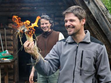 Zwei Männer entzünden Feuer in einem mit Gras befüllten Stück Birkenrinde