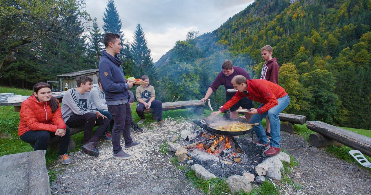 Junge Menschen sitzen um ein Lagerfeuer auf dem in einer Pfanne Essen gekocht wird